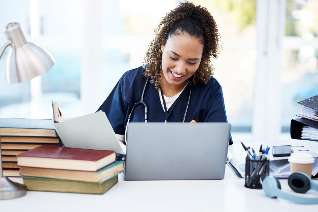 Nurse in scrubs smiling and working on a laptop.